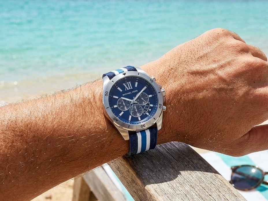 Man wearing a Michael Kors chronograph watch with blue dial and navy-and-white striped band, resting his arm on a beach chair with turquoise ocean and palm trees in the background.