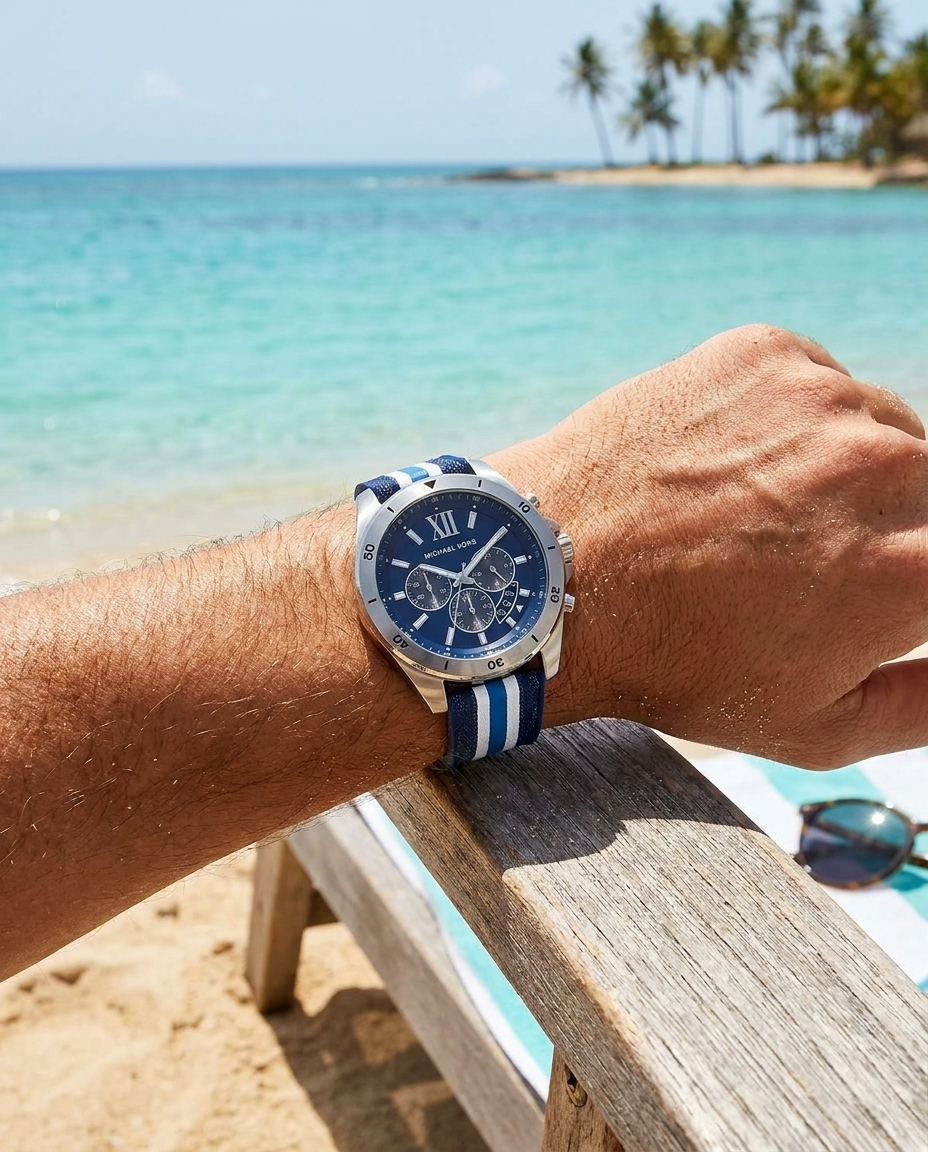 Man wearing a Michael Kors chronograph watch with blue dial and navy-and-white striped band, resting his arm on a beach chair with turquoise ocean and palm trees in the background.